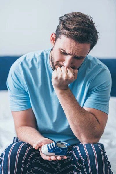 Grieving father sitting on bed, covering mouth with hand and holding baby shoe at home — Stock Photo