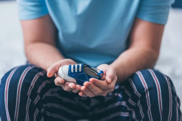 Cropped view of man sitting on bed and holding baby shoe — Stock Photo