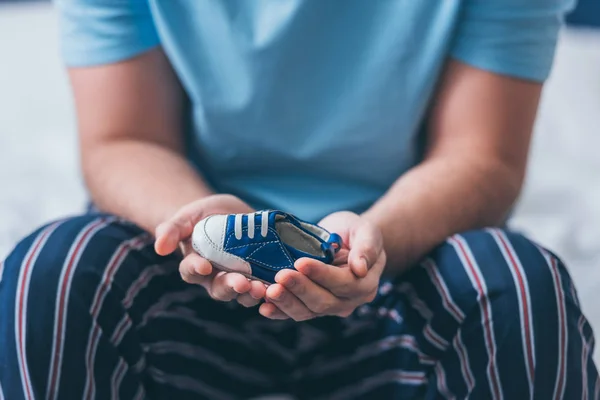 Vista ritagliato di uomo seduto sul letto e tenendo scarpa bambino — Foto stock