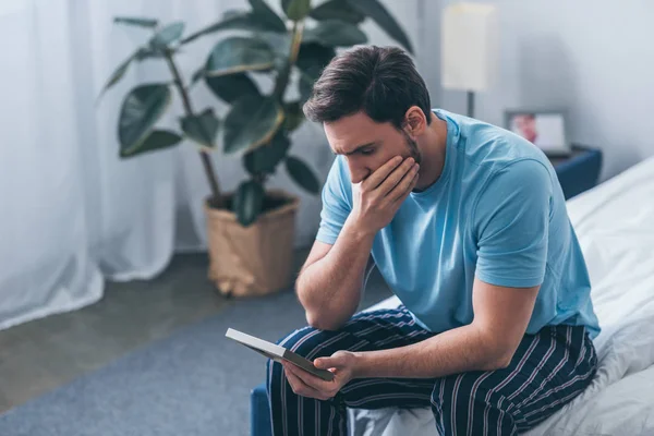 Aufgebrachter Mann sitzt auf Bett, deckt Mund zu und hält Fotorahmen zu Hause — Stockfoto