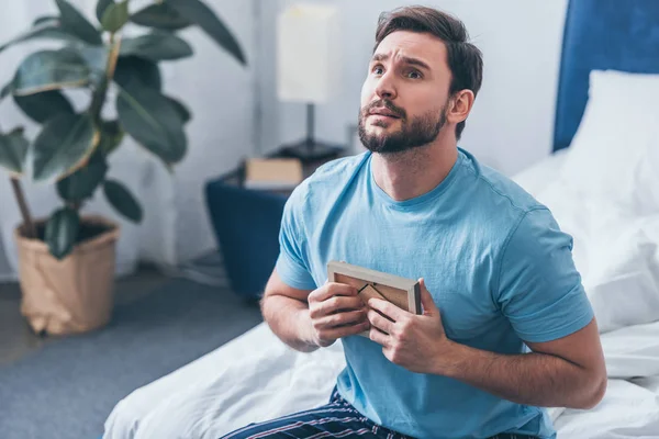 Grieving man sitting on bed, looking up and holding photo frame at home — Stock Photo