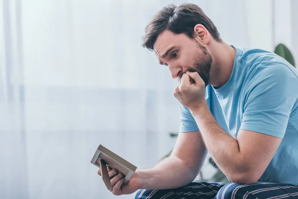 Uomo depresso seduto, che copre la bocca con la mano e guardando cornice a casa — Foto stock