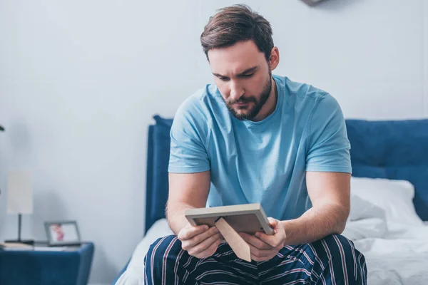 Grieving man sitting on bed and looking at photo frame at home with copy space — Stock Photo