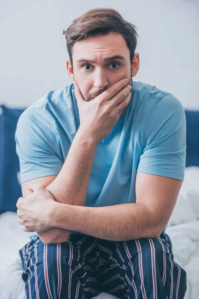 Homme stressé assis sur le lit, regardant la caméra et couvrant la bouche avec la main à la maison — Photo de stock