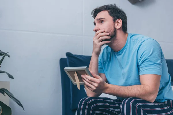 Uomo sconvolto seduto sul letto, che copre la bocca e tiene la cornice a casa — Foto stock