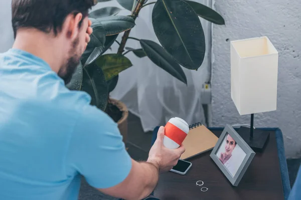 Man photo holding funeral urn, covering face with hand and grieving near picture of woman and wedding rings — Stock Photo