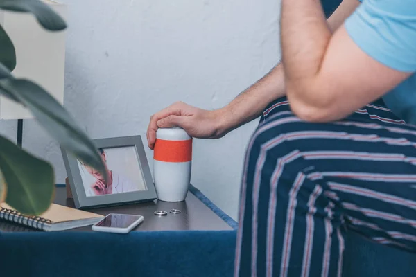 Cropped view of man holding funeral urn near picture of woman in frame and wedding rings on table — Stock Photo