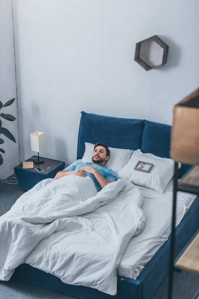 Depressed man lying in bed near photo frame with picture of woman on pillow at home — Stock Photo