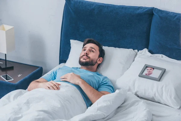 Hombre deprimido acostado en la cama cerca del marco de la foto con la imagen de la mujer en la almohada en casa - foto de stock