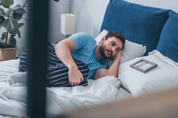 Selektiver Fokus eines traurigen Mannes, der im Bett neben einem Bild der Frau auf dem Kopfkissen zu Hause liegt — Stockfoto