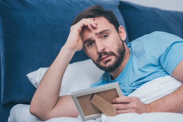 Upset man lying in bed, looking at camera and holding photo frame at home — Stock Photo