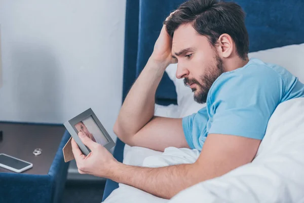 Hombre molesto acostado en la cama, tocando la cabeza y sosteniendo el marco de fotos en casa - foto de stock