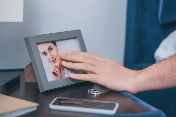 Vista recortada de hombre tocando marco de foto con foto de mujer cerca de anillos de boda y teléfono inteligente - foto de stock