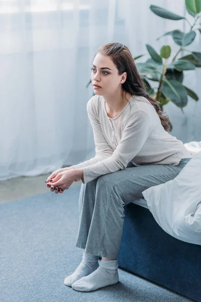 Сумна жінка в піжамі зі складеними руками сидить на ліжку вдома — стокове фото