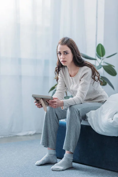 Mujer afligida sentada en la cama, mirando a la cámara y sosteniendo el marco de la foto en casa — Stock Photo