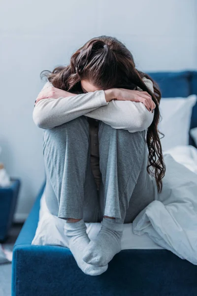 Femme déprimée avec tête baissée étreignant les genoux et assise sur le lit à la maison — Photo de stock