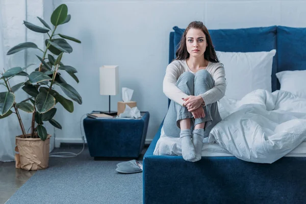 Bouleversé femme étreignant les genoux, regardant la caméra et assis sur le lit à la maison — Photo de stock