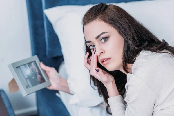 Beautiful upset woman holding picture frame, crying and looking at camera at home — Stock Photo