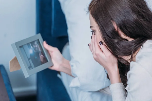 Femme bouleversée couchée au lit, pleurant et regardant le cadre avec la photo de l'homme à la maison — Photo de stock