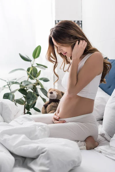 Brunette pregnant woman sitting on bed and touching tummy — Stock Photo