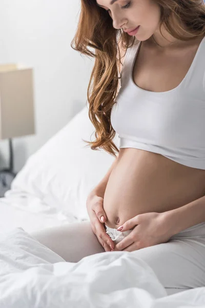 Довгошерста вагітна жінка торкається живота сидячи в ліжку — стокове фото