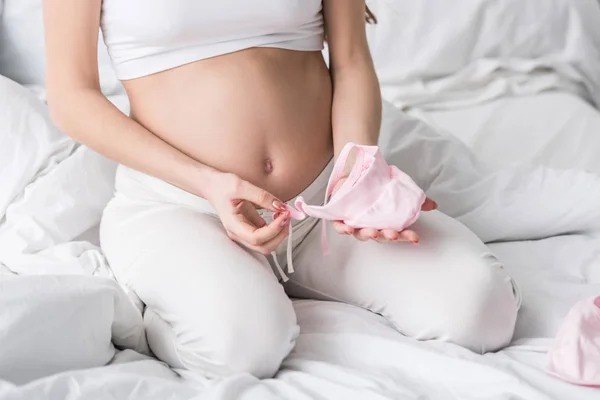Vista ritagliata della donna incinta che tiene i vestiti rosa del bambino mentre si siede sul letto — Foto stock