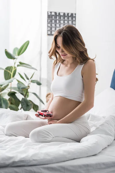 Sourire femme enceinte toucher le ventre et tenant voiture jouet rouge — Photo de stock