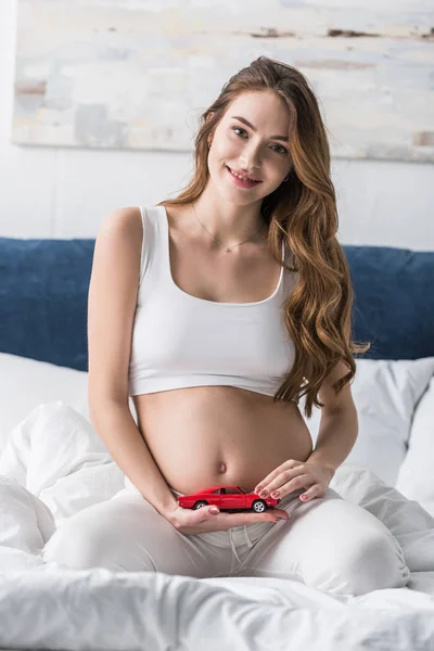 Mujer embarazada dichosa sentada en la cama y sosteniendo el coche de juguete rojo - foto de stock