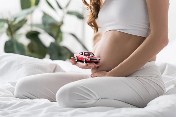 Vista parcial de la mujer embarazada sosteniendo coche de juguete rojo - foto de stock