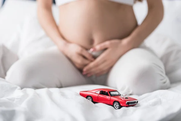 Vista parcial de la mujer embarazada sentada en la cama con coche de juguete rojo - foto de stock