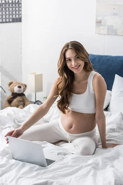 Sonriendo chica embarazada sentada en la cama con el ordenador portátil y mirando a la cámara - foto de stock