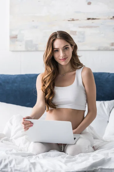 Hermosa mujer embarazada usando el ordenador portátil mientras está sentado en la cama - foto de stock