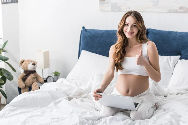 Sonriendo mujer embarazada usando el ordenador portátil en la cama y mostrando el pulgar hacia arriba - foto de stock