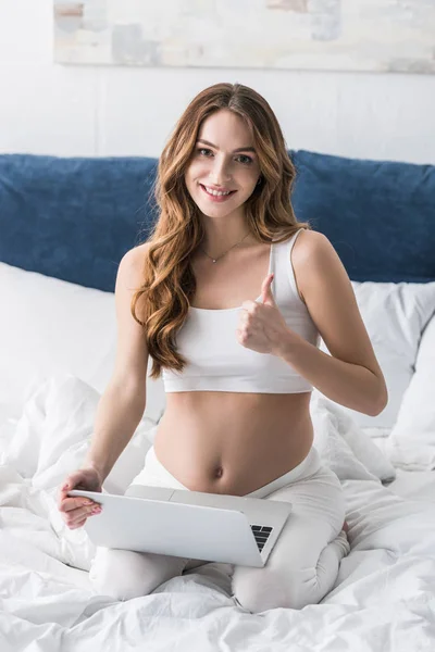 Curly pregnant woman using laptop in bed and showing thumb up — Stock Photo