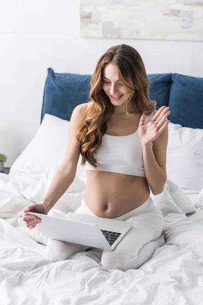 Mujer embarazada rizado utilizando el ordenador portátil y la mano ondulante - foto de stock