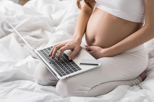Vista parcial de la mujer embarazada sentada en la cama con ordenador portátil y tarjeta de crédito - foto de stock