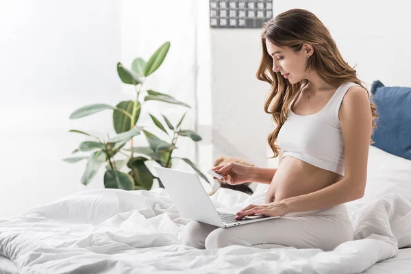 Hermosa mujer embarazada sentada en la cama con computadora portátil y tarjeta de crédito - foto de stock