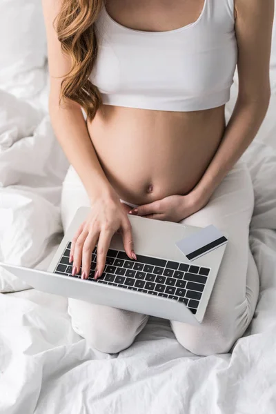 Vista parcial de la mujer embarazada sentada en la cama con ordenador portátil y tarjeta de crédito - foto de stock