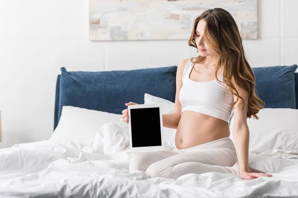 Lockige schwangere Frau sitzt auf dem Bett und hält ein digitales Tablet mit leerem Bildschirm — Stockfoto