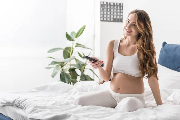 Улыбающаяся беременная девушка сидит на кровати и держит пульт от телевизора — стоковое фото