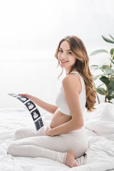 Happy pregnant woman sitting on bed and holding ultrasound scans — Stock Photo