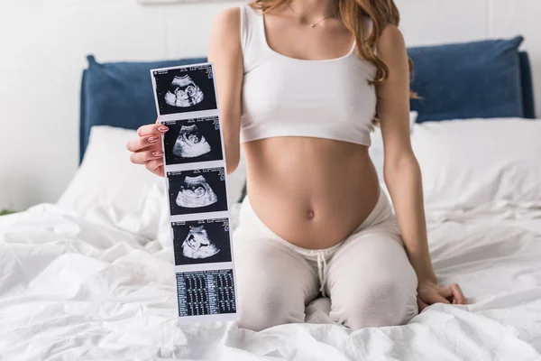 Обрезанный вид беременной женщины, сидящей на кровати и показывающей ультразвуковое сканирование — стоковое фото
