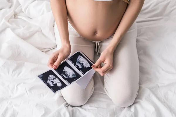 Partial view of pregnant woman sitting on bed with ultrasound scans — Stock Photo