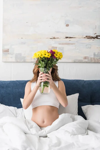 Pregnant woman sitting in bed and holding vase with flowers — Stock Photo