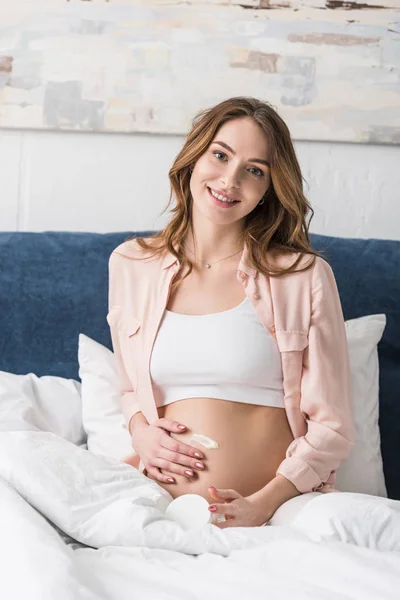 Happy pregnant woman sitting on bed and applying cream on belly — Stock Photo