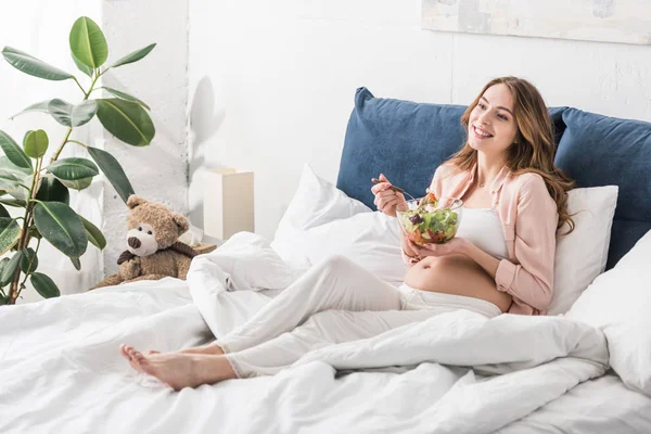 Mujer embarazada inspirada comiendo ensalada en la cama - foto de stock