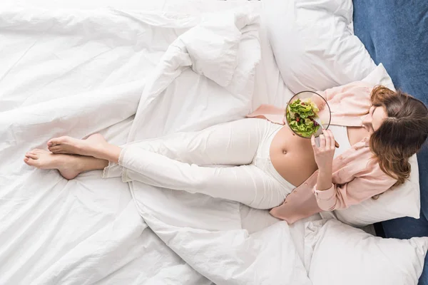 Vista superior de la mujer embarazada comiendo ensalada en la cama - foto de stock