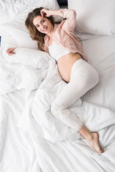 Вид сверху довольной беременной женщины, лежащей в постели с улыбкой — стоковое фото