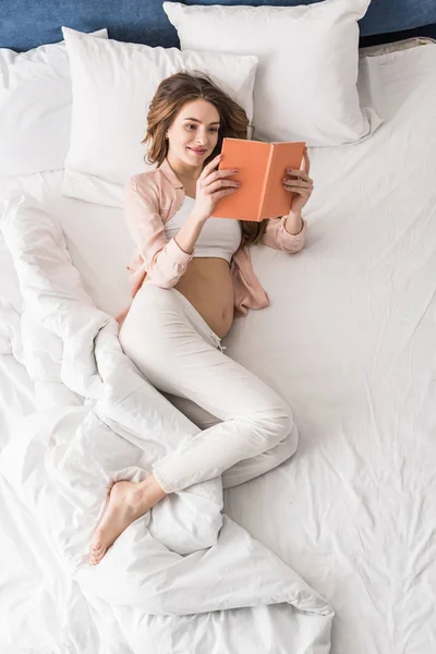Vista superior de la mujer embarazada sonriente leyendo libro en la cama - foto de stock