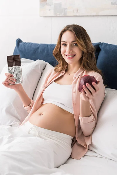 Donna incinta sorridente con barretta di cioccolato e mela a letto — Foto stock